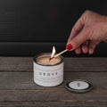 Hand lighting the Mini Grove orange blossom candle by backhouse with a match against a dark background.