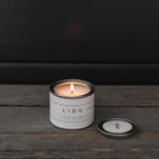 Mini Lido sandalwood candle by backhouse burning on a wood surface with the metal lid placed beside it against a dark background.