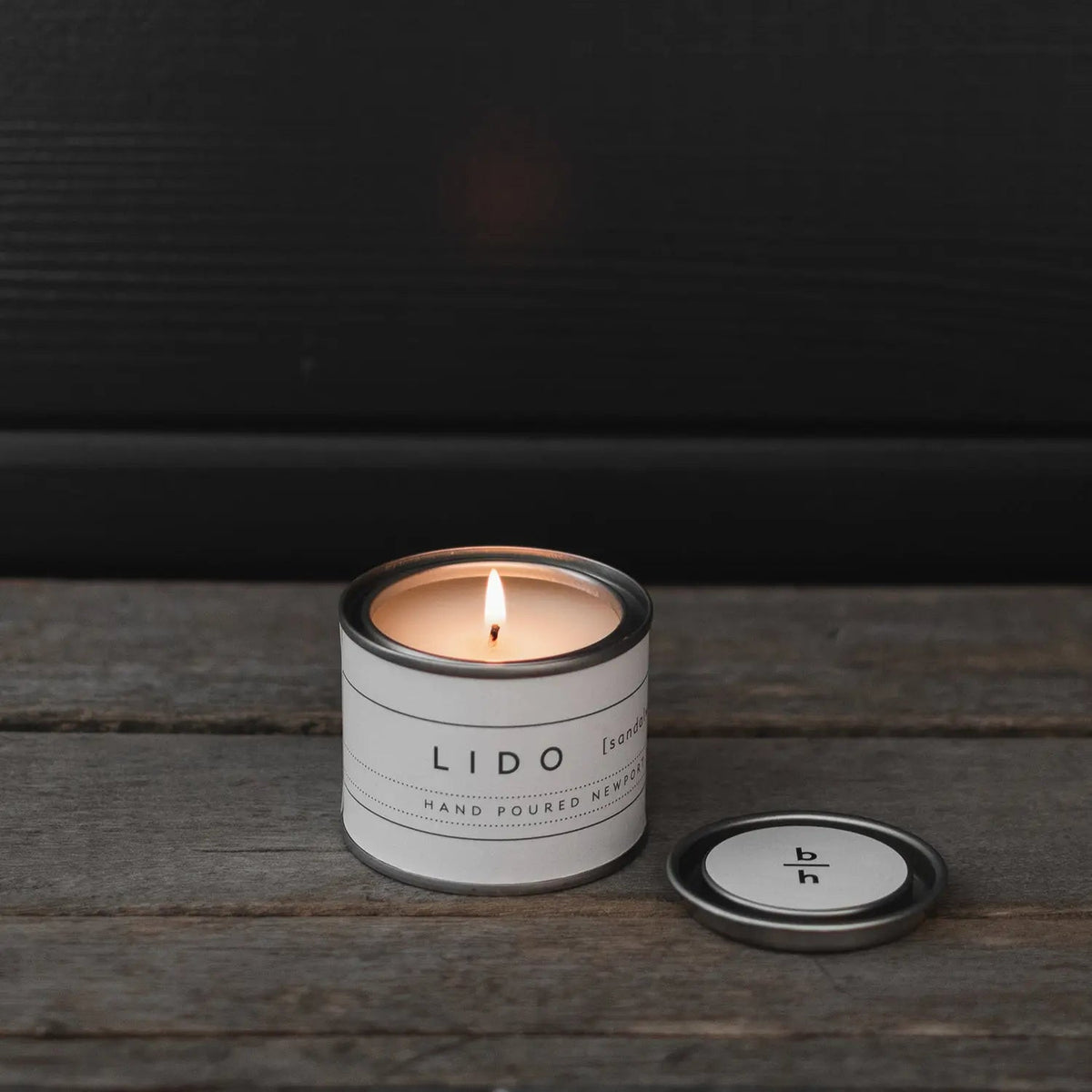 Mini Lido sandalwood candle by backhouse burning on a wood surface with the metal lid placed beside it against a dark background.
