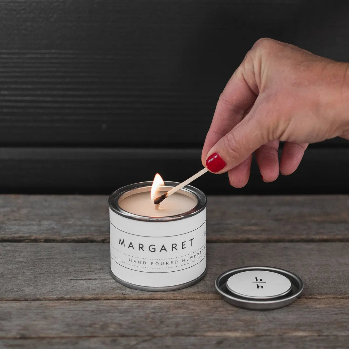 Hand lighting the Mini Margaret currant candle by backhouse with a match on a wood surface against a dark background.
