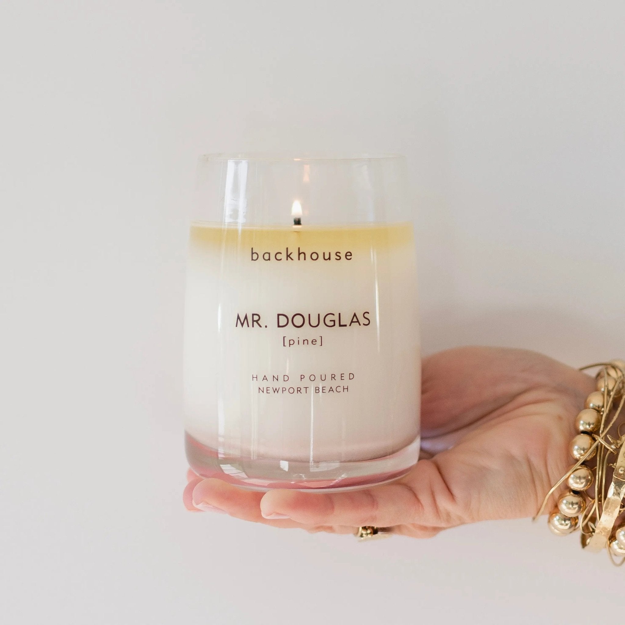 Mr. Douglas pine candle by backhouse lit and held in hand against a white background, shown in its classic glass vessel.