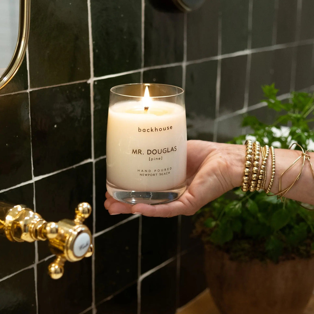 Mr. Douglas pine candle by backhouse lit and held against a dark green tile background in a bathroom setting.
