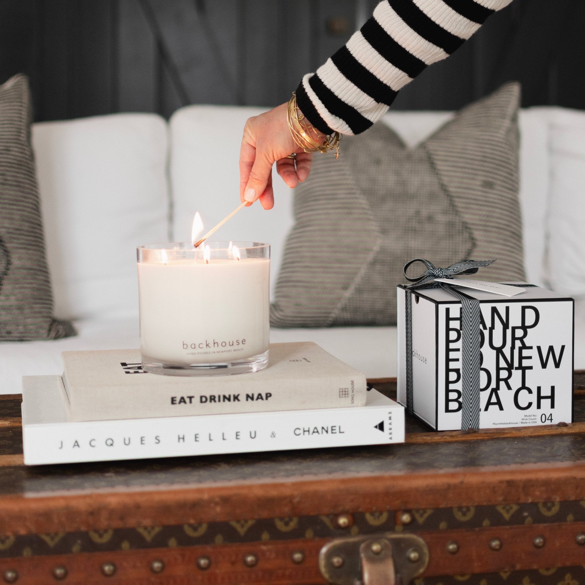 A hand wearing gold bracelets and a black and white striped sweater lighting a white three wick backhouse candle sitting on top of coffee table books including Eat Drink Nap and Jacques Helleu Chanel, next to a backhouse Newport Beach gift box with a black ribbon bow.