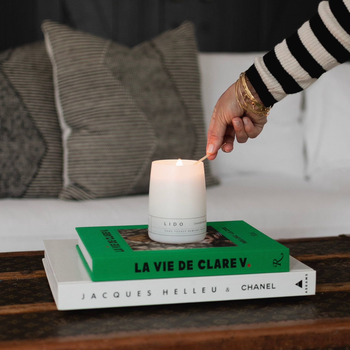 A white LIDO scented candle being lit with a match, sitting atop stacked fashion books including La Vie de Clare V. and Jacques Helleu & Chanel on a vintage trunk Newport Beach
