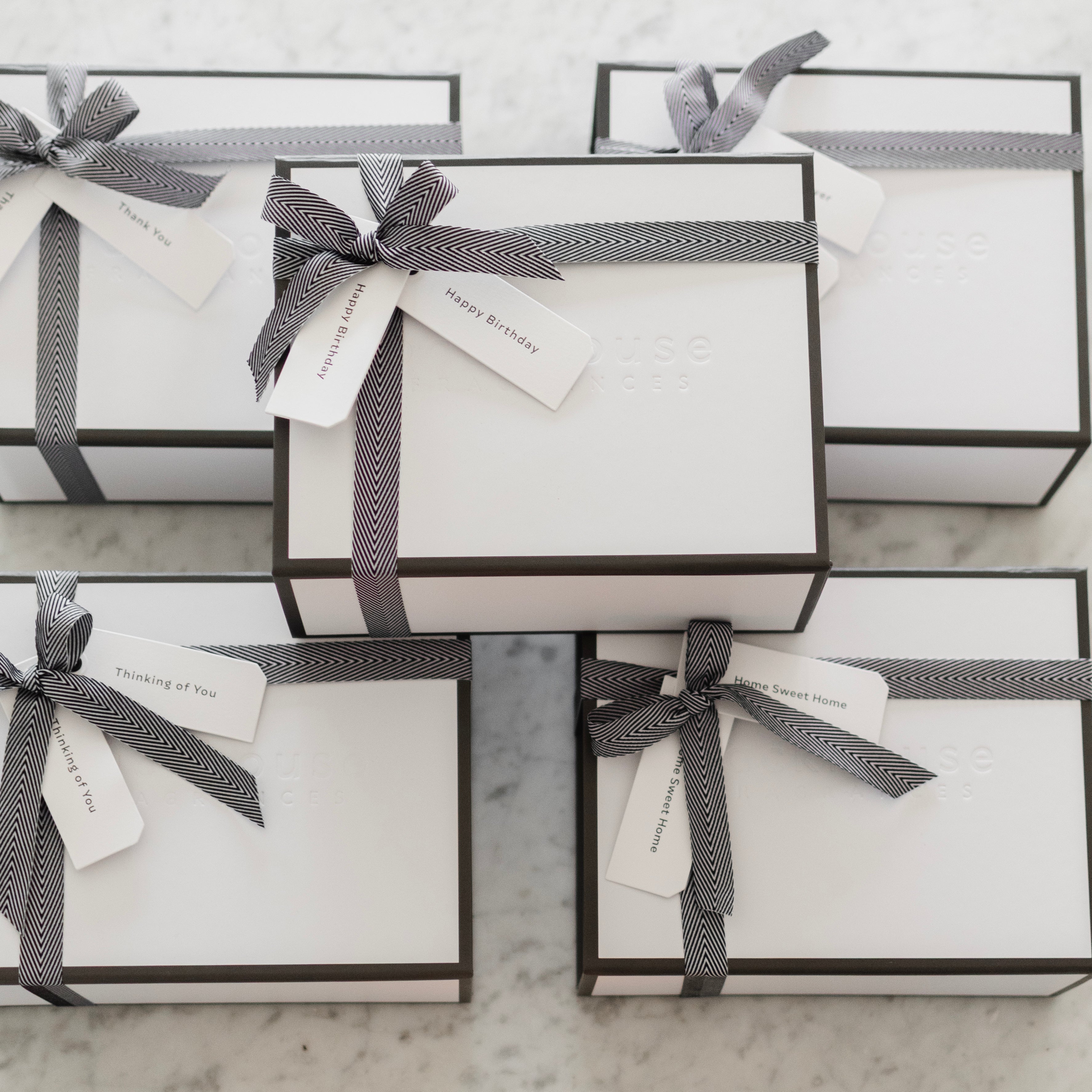 Several elegant white gift boxes with black trim and black herringbone ribbon bows, tagged with Happy Birthday, Thinking of You, Thank You, and Home Sweet Home labels, arranged on a marble surface with a small candle nearby.
