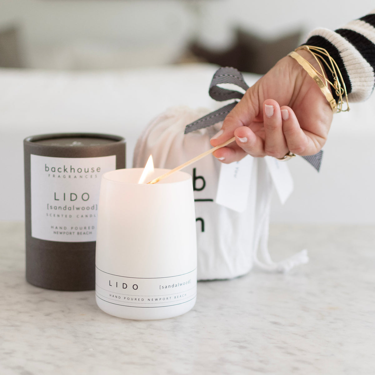 A hand lighting a Backhouse Fragrances Lido sandalwood scented candle in a white matte vessel on a marble surface, with the Grey cylindrical packaging box and a white gift bag visible in the background.