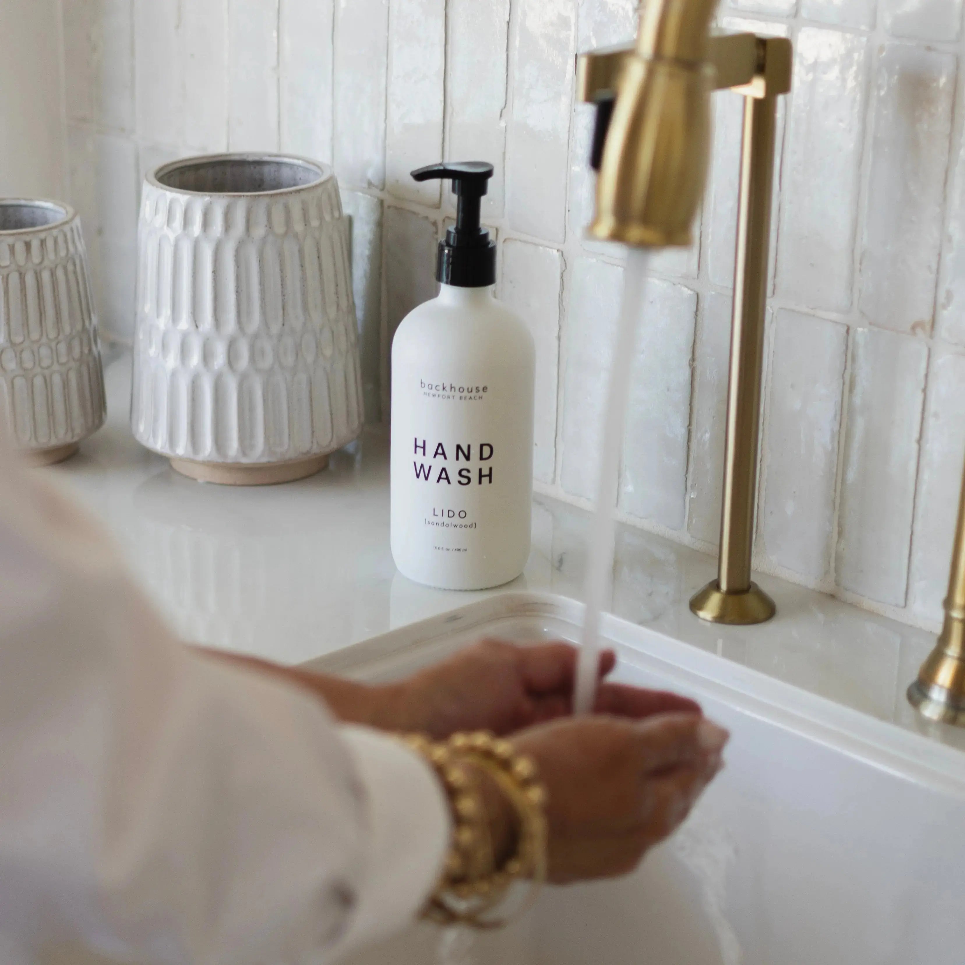 Hands pouring the Lido sandalwood 32oz hand wash refill by backhouse into a glass bottle at a sink.