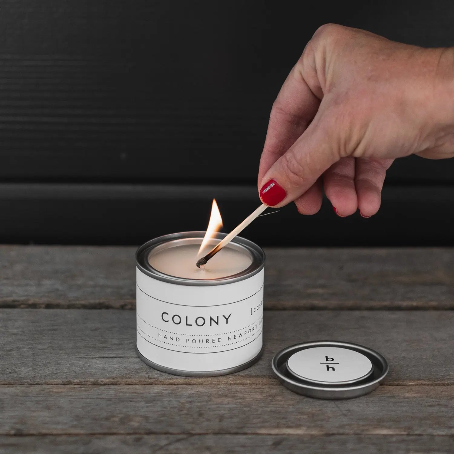 Hand lighting the Mini Colony cassis candle by backhouse with a match on a wood surface against a dark background.
