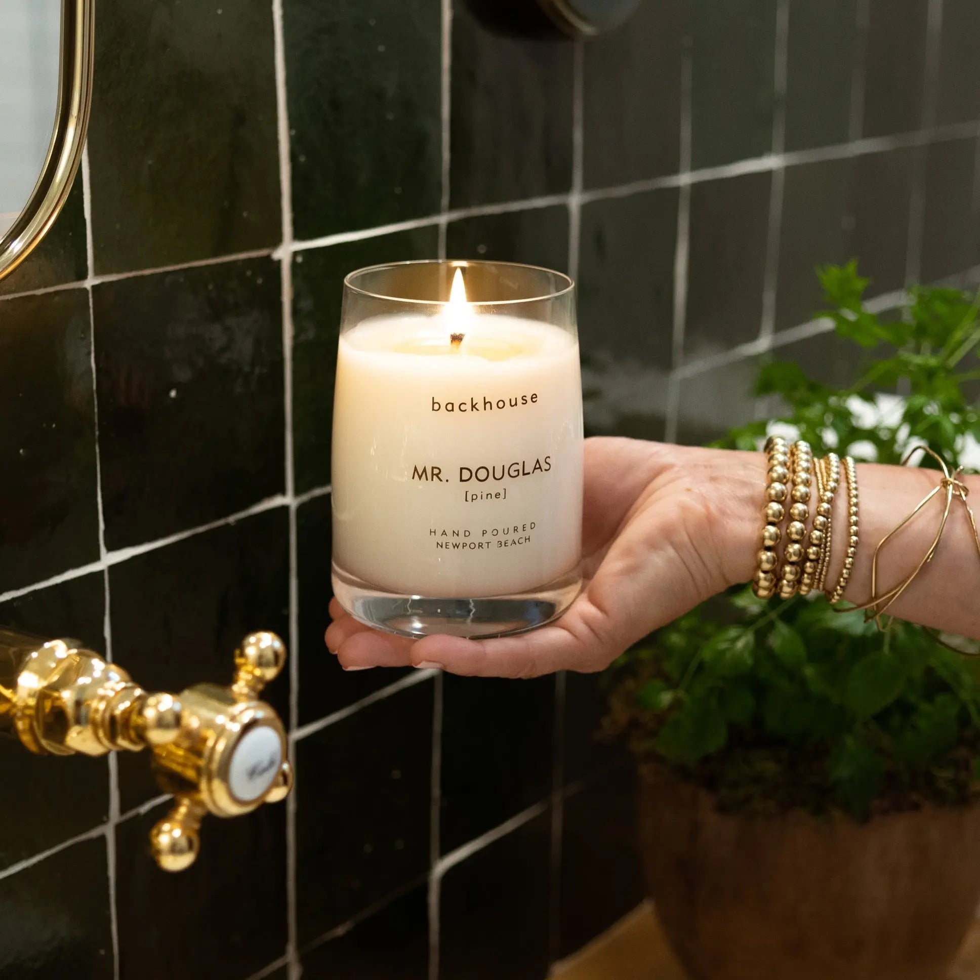 Mr. Douglas pine candle by backhouse lit and held against a dark green tile background in a bathroom setting.