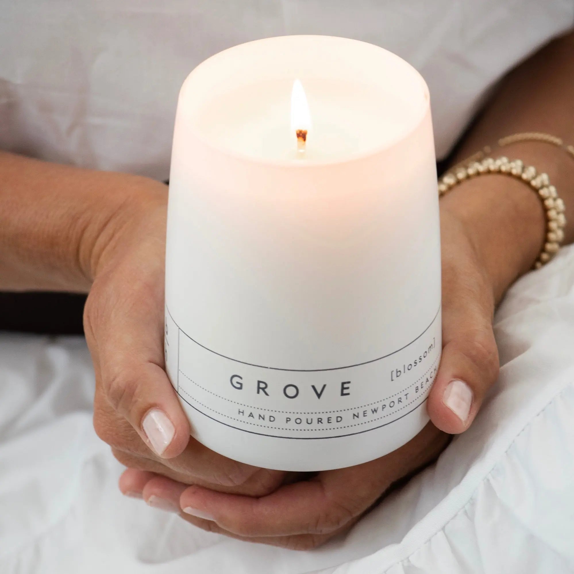 Grove orange blossom candle by backhouse burning while being held in both hands in a soft neutral setting.